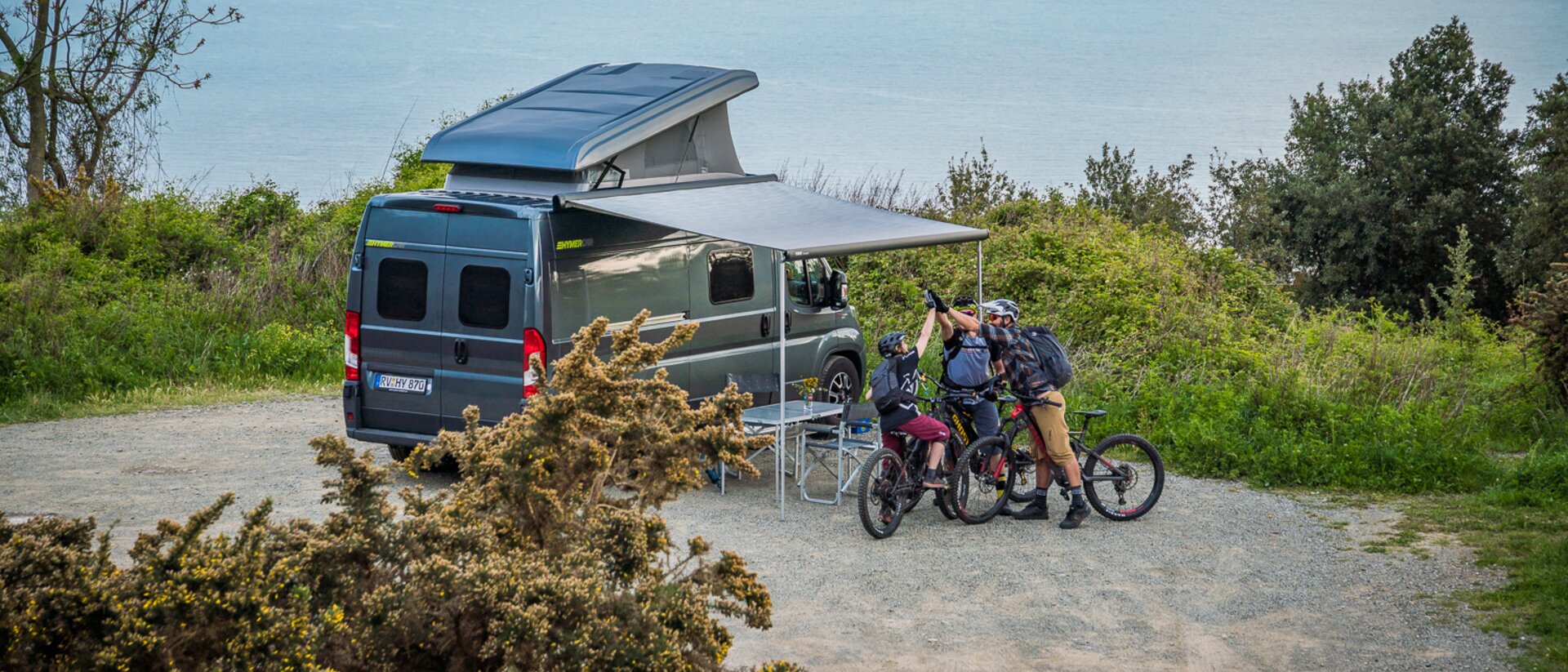 Campingsituasjon ved kysten med HYMER Camper Van med utfoldet markise / syklist foran kjøretøyet.